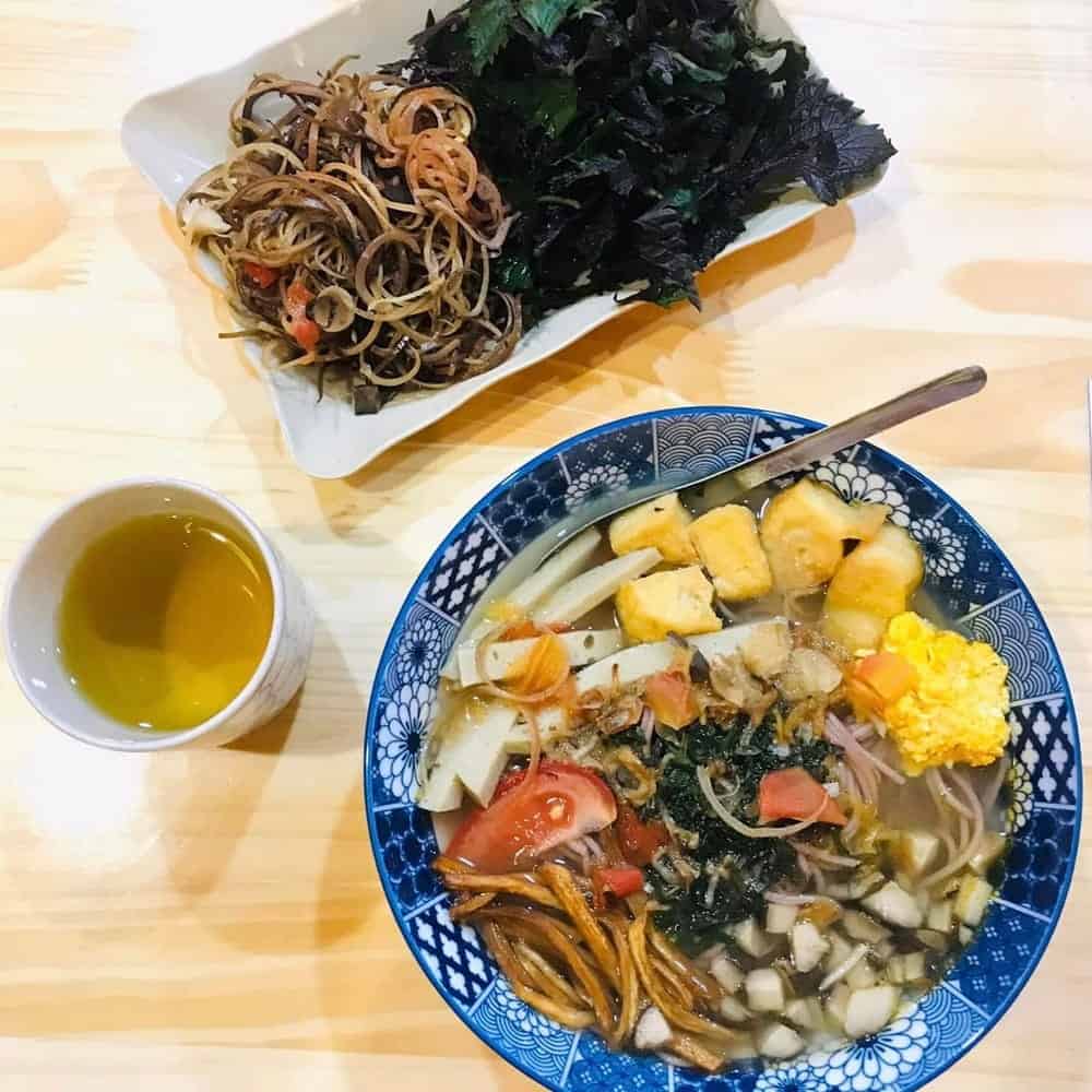 large bowl of vegan noodle soup with veggies next to a small rectangle plate with noodles and green veggies and a coffee at hem in hanoi