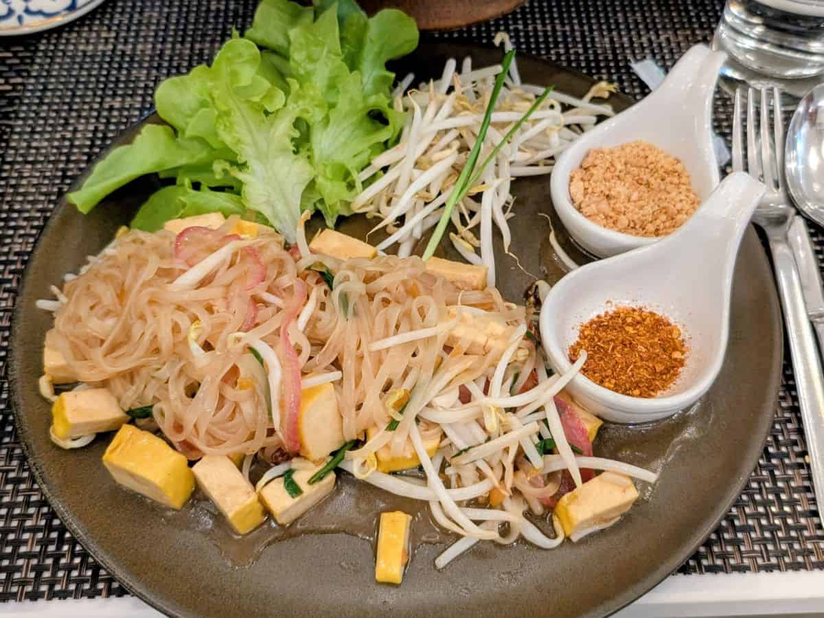 a large black plate with vegan pad thai with chunks of tofu next to small bowls of chili and peanuts in thailand