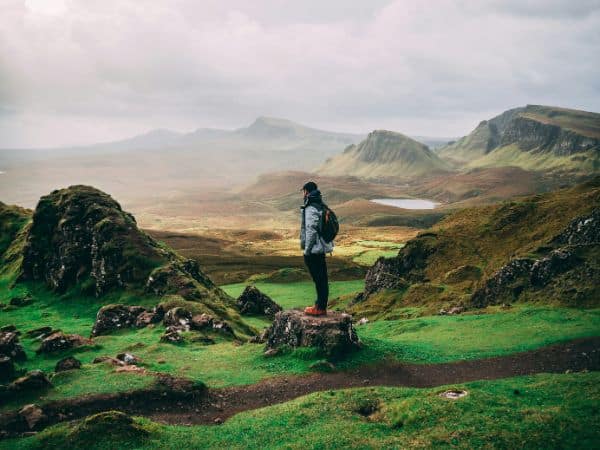 a single hiker standing on a path amongst large rocks and green hills on a misty day in scotland