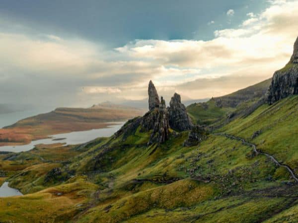 clouds rolling over a lush green mountain with two large rock formations surrounded by water in scotland