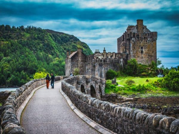 two people walking a stone path to a small castle surrounded by lush greenery in scotland
