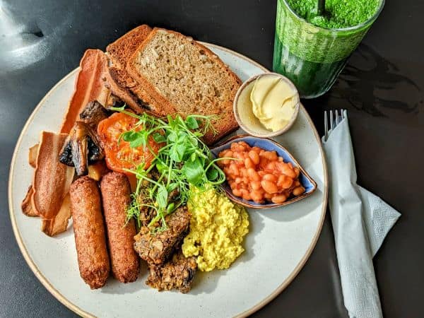 large white plate with vegan sausage, tofu, mushrooms, tomato, and toast with a green juice in scotland