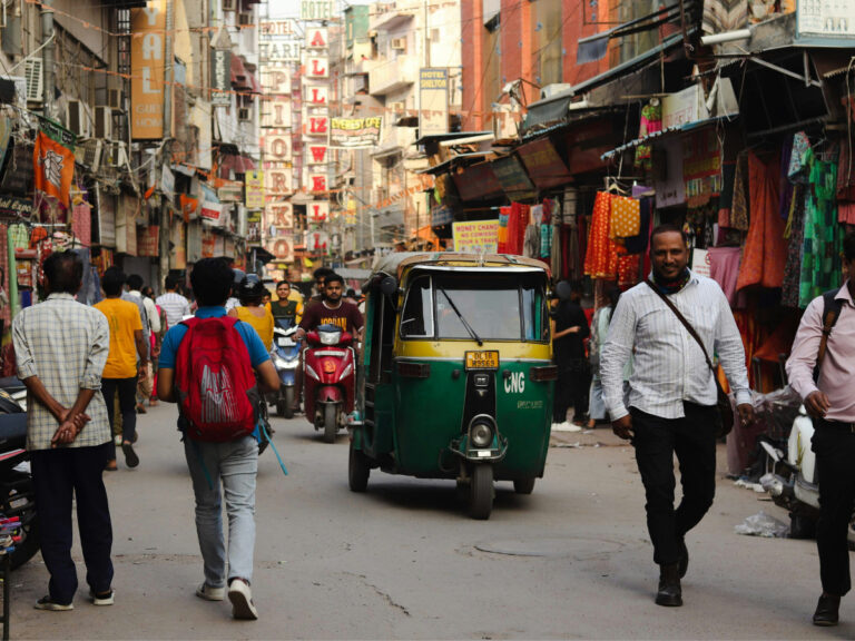 Green and yellow auto rickshaw driving through a bustling market street in Delhi, India, surrounded by shops, pedestrians, and colorful storefronts.