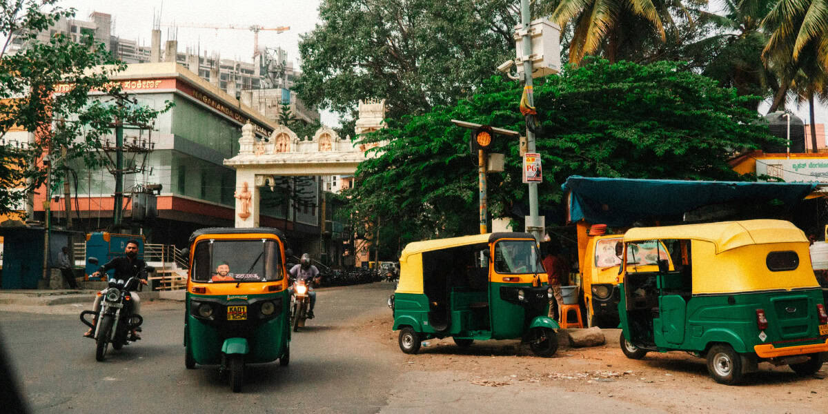 Green and yellow auto rickshaw driving through a bustling market street in Delhi, India, surrounded by shops, pedestrians, and colorful storefronts.