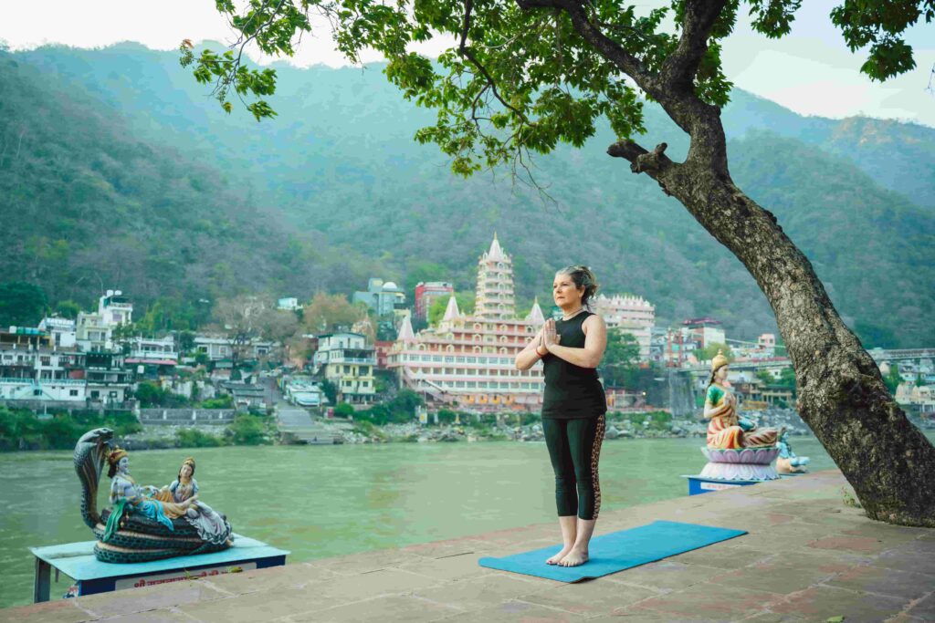 Traveler practicing yoga beside the Ganges River in Rishikesh, India, with temples and forested hills in the background.