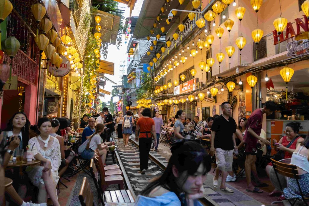 People dining at crowded outdoor restaurants along Hanoi's Train Street, illuminated by yellow lanterns.